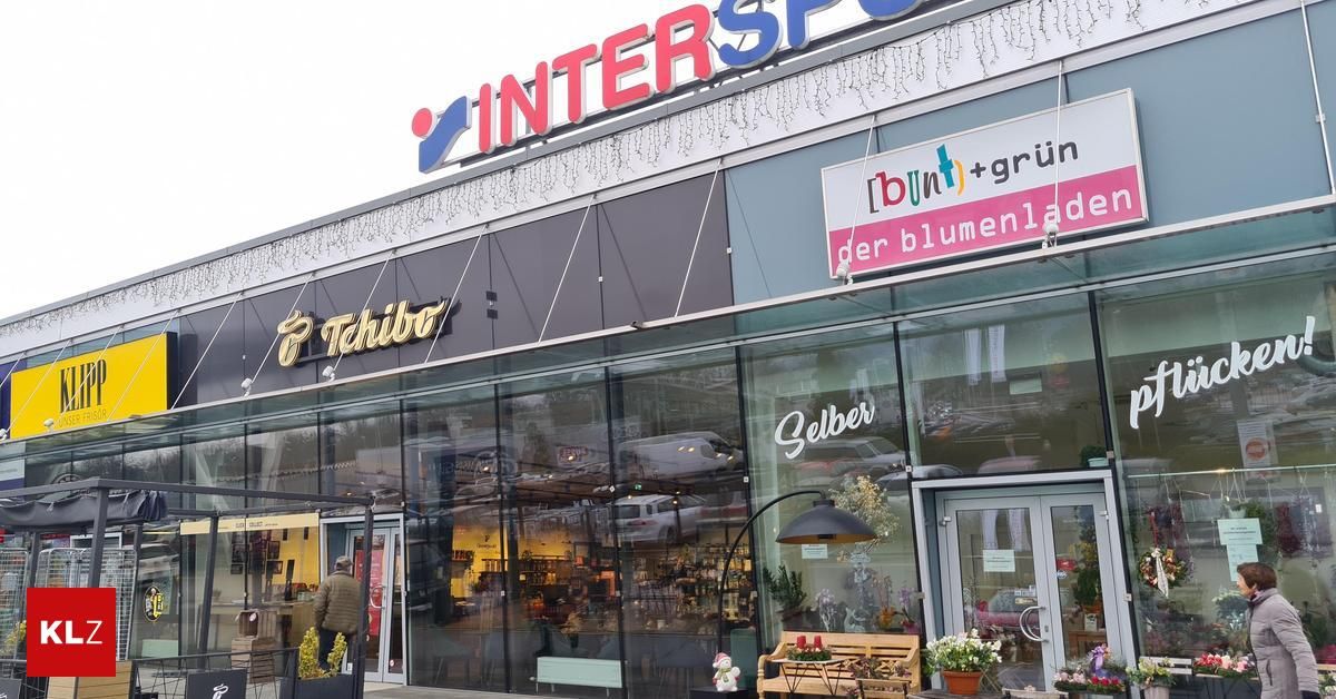Die Fassade des Intersport-Geschäfts, einem Sportartikelladen, zeigt durch Glaswände verschiedene Sportgeräte. Ein Schild mit der Aufschrift 'der blumenladen' hängt über dem Eingang, und eine Bank mit einem Topf steht draußen.