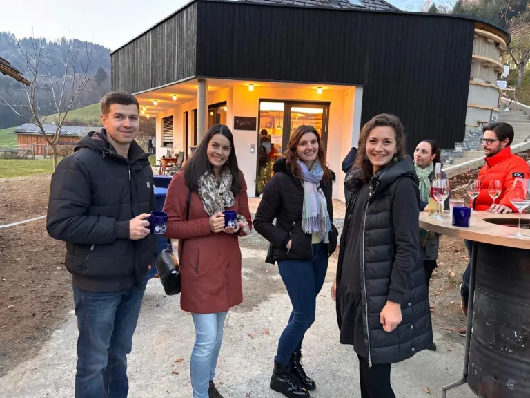 Fünf Personen stehen vor einem modernen Gebäude, lächeln und halten Tassen. Zwei Frauen tragen Schals, und zwei andere haben die Hände in den Taschen. Ein Tisch mit Weingläsern ist in der Nähe.