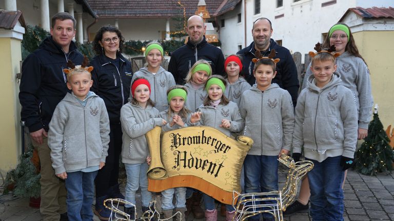 Eine Gruppe von Erwachsenen und Kindern steht vor einem Gebäude. Sie tragen alle graue Hoodies und Stirnbänder. Die Kinder halten ein Holzschild mit der Aufschrift Bromberger Advent.