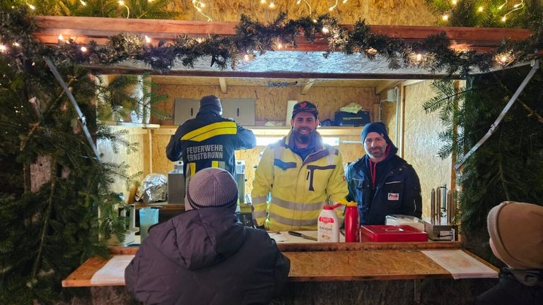 Drei Männer in Jacken stehen in einer Kabine mit Weihnachtsdekoration. Der Mann in der Mitte lächelt. Auf dem Tresen befinden sich eine Flasche, ein Becher und ein Behälter.