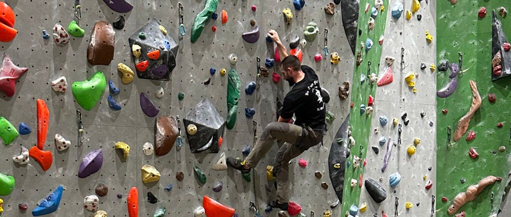 Ein Mann in einem schwarzen Shirt und Hose klettert an einer Wand mit vielen unterschiedlich gefärbten und geformten Felsen.