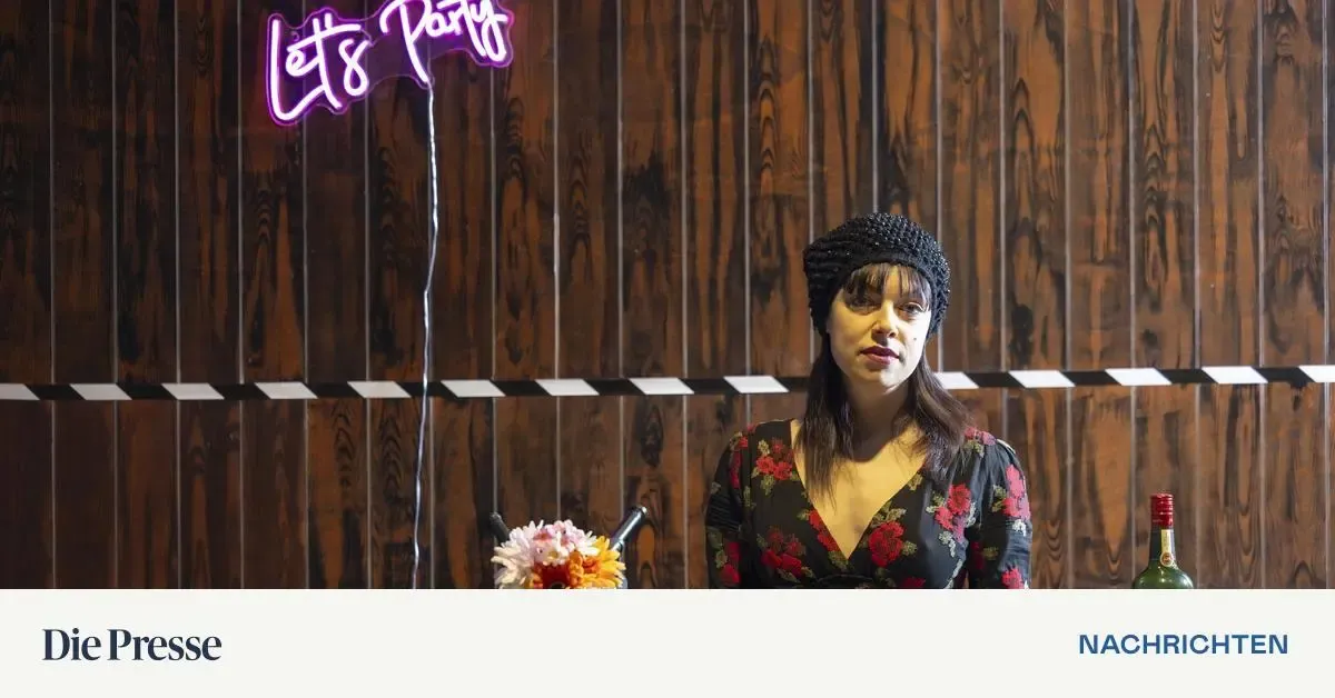 Eine Frau in einem Blumenkleid und einer Mütze steht vor einer Holzwand mit einem Neonschild 'Let's Party' und einer Blumenarrangement.