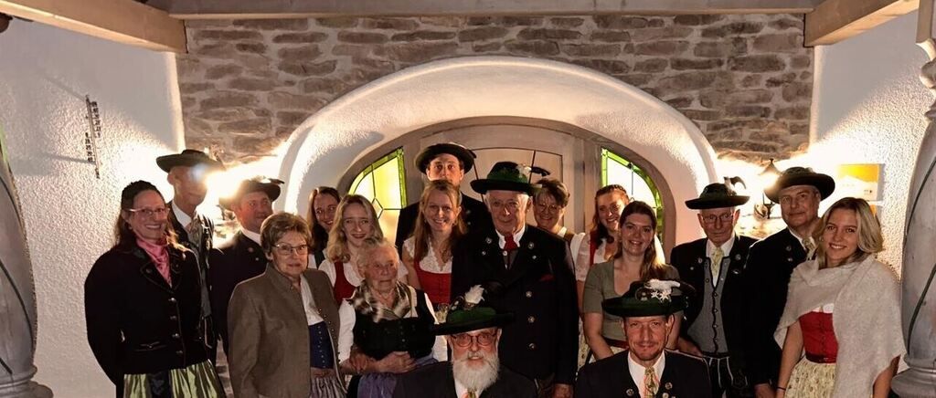 Eine Gruppe von Menschen in traditioneller bayerischer Kleidung posiert für ein Foto in einem Steinzimmer. Sie lächeln und stehen eng beieinander.