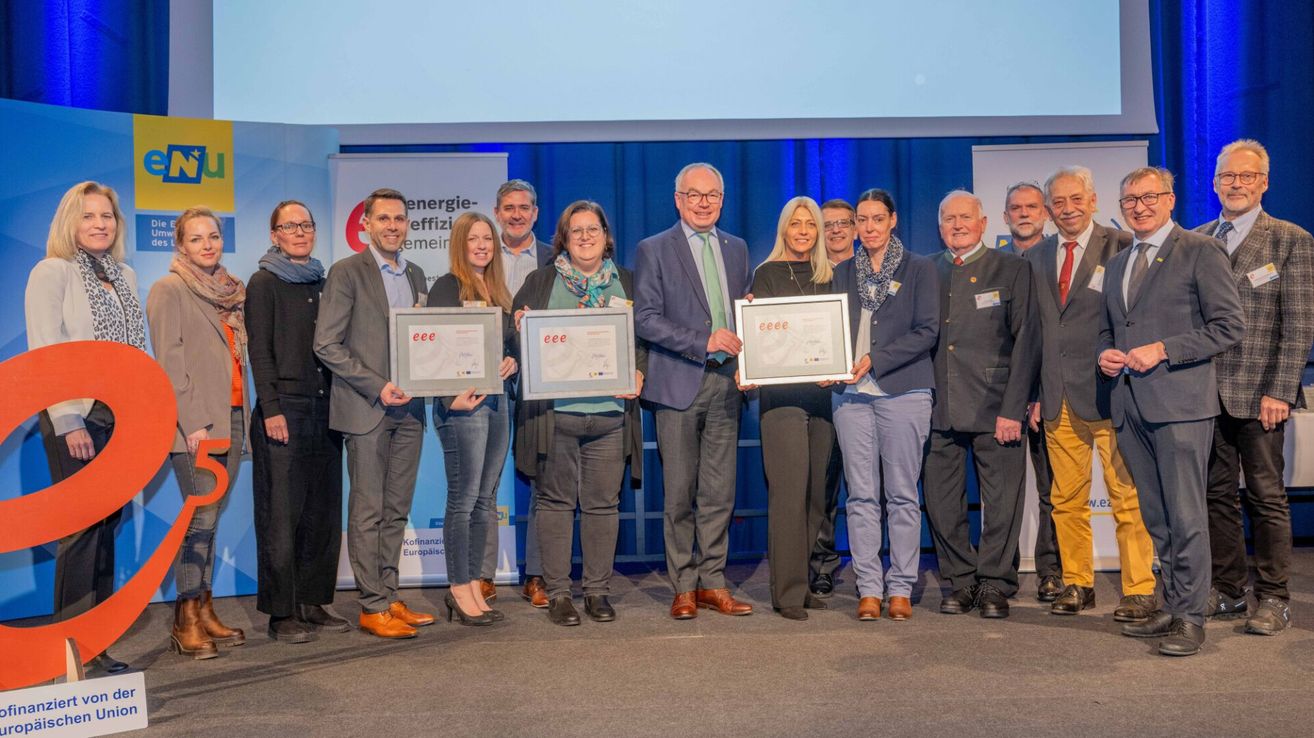 Eine Gruppe von Personen in formeller Kleidung, die in einer Reihe stehen und Urkunden halten, mit einem Hintergrund von Bannern und einer Bühne.