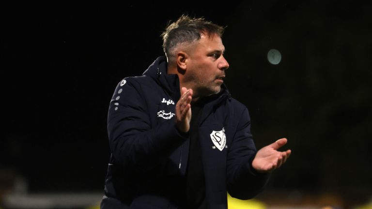 Ein Mann mit einem schwarzen Jacken und einem Logo auf der linken Brust klatscht auf einem Feld mit den Händen.