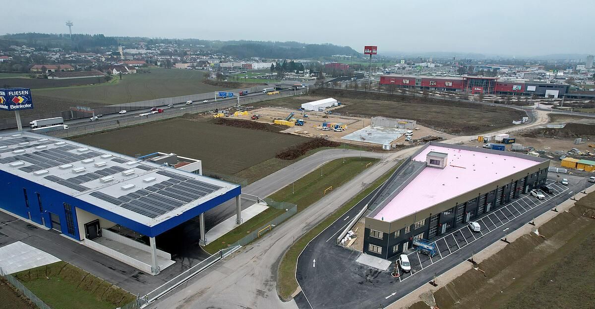 Ein Luftbild eines Baugeländes mit verschiedenen Fahrzeugen und Gebäuden im Bau, einer Autobahn mit Autos und einer Stadtlandschaft in der Ferne.