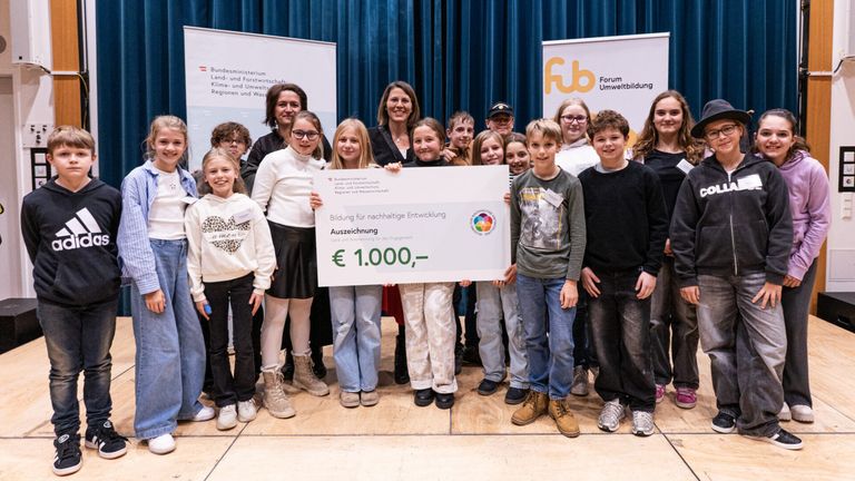 Eine Gruppe von Kindern steht vor einer Bühne und hält einen großen Scheck über 1.000 Euro. Der Scheck trägt die Aufschrift 'Bildung für nachhaltige Entwicklung' und wird von einem Mädchen gehalten. Die Gruppe scheint eine Anerkennung oder Auszeichnung zu feiern.