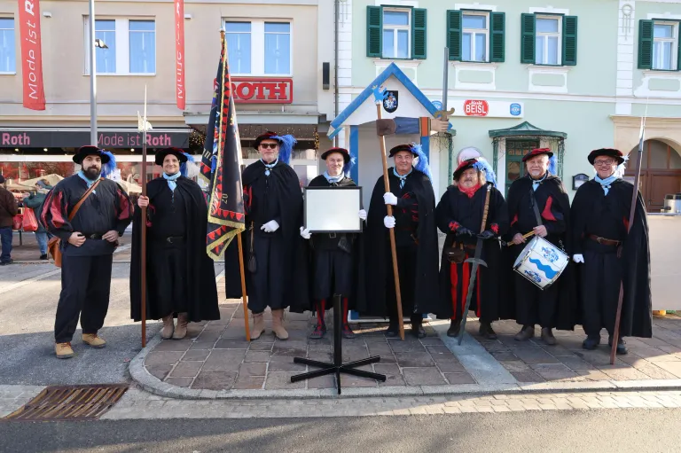 Eine Gruppe von Menschen in traditioneller Kleidung, einschließlich Hüten, Handschuhen und Stiefeln, steht in einer Reihe und hält eine Flagge, Schwerter und eine Trommel. Sie stehen vor einem Gebäude mit einem Schild, auf dem 'Roth' steht.
