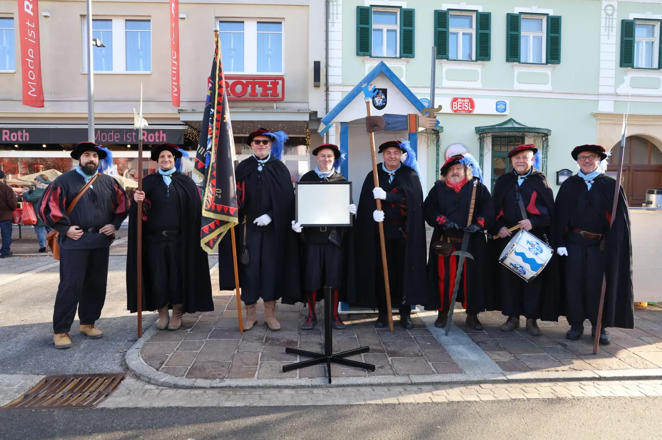 Eine Gruppe von Menschen in traditioneller Kleidung, einschließlich Hüten, Handschuhen und Stiefeln, steht in einer Reihe und hält eine Flagge, Schwerter und eine Trommel. Sie stehen vor einem Gebäude mit einem Schild, auf dem 'Roth' steht.