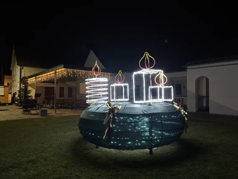 Ein großer beleuchteter Kranz mit LED-Kerzen steht bei Nacht auf einem Rasen. Dahinter ein Gebäude mit Bänken und einem Dach mit Lichterketten.
