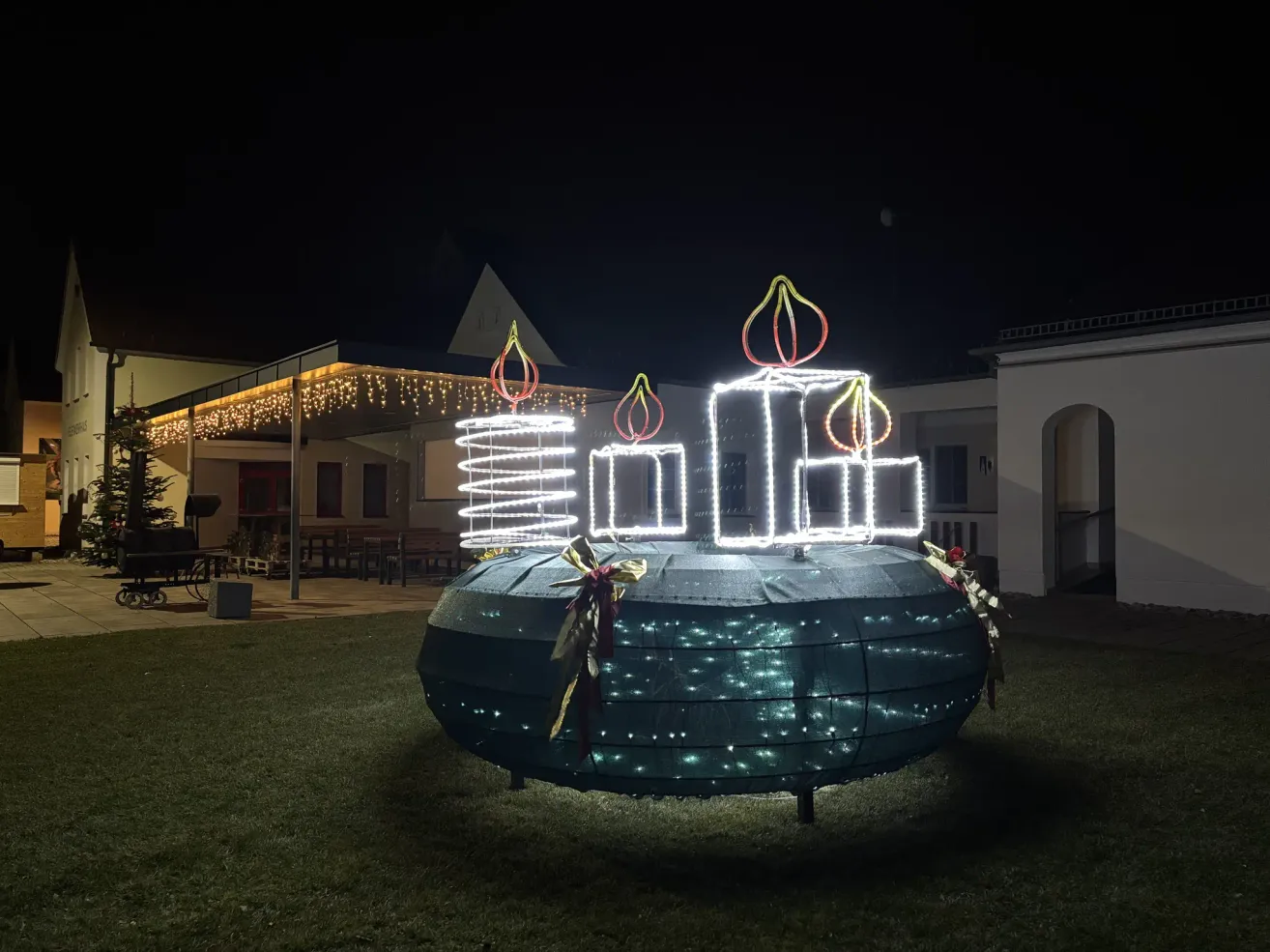 Ein großer beleuchteter Kranz mit LED-Kerzen steht bei Nacht auf einem Rasen. Dahinter ein Gebäude mit Bänken und einem Dach mit Lichterketten.