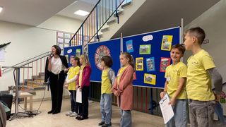Eine Frau steht mit einem Mikrofon und spricht zu einer Gruppe von Kindern in gelben Shirts, die vor einem blauen Bulletin-Board mit Bildern stehen.