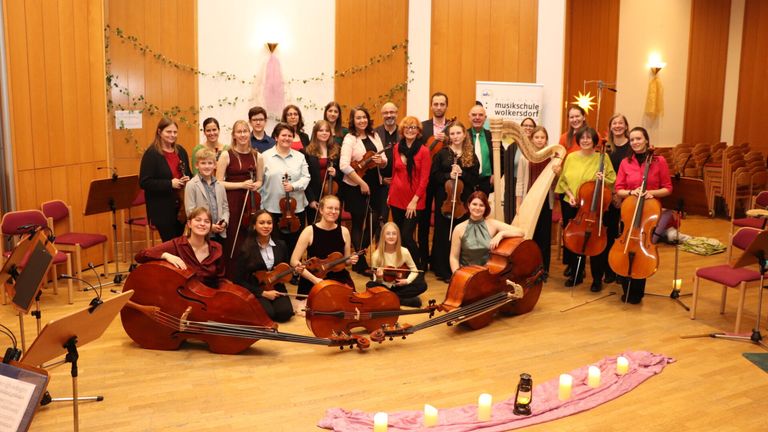 Eine Gruppe von Musikern posiert mit ihren Instrumenten auf einem Holzboden. Einige sitzen auf dem Boden, während andere hinter ihnen stehen. Kerzen sind in einem Kreis um sie herum angeordnet.