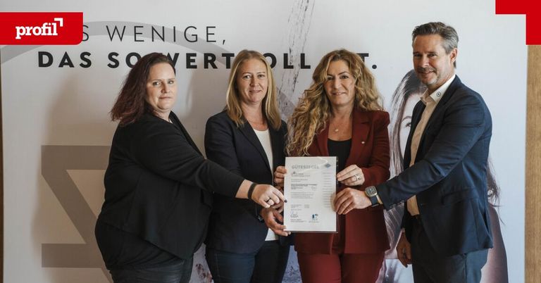 Vier Personen halten ein Zertifikat und posieren für ein Foto. Eine Frau in Rot hält einen Stift. Die Frau in Schwarz lächelt. Hinter ihnen ist ein großer Banner mit Text.