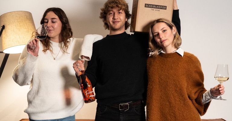 Drei Menschen stehen in einem Raum zusammen. Ein Mann in der Mitte hält eine Flasche Wein, während eine Frau links aus einem Glas trinkt. Eine Frau rechts lächelt. Sie stehen vor einer weißen Wand.