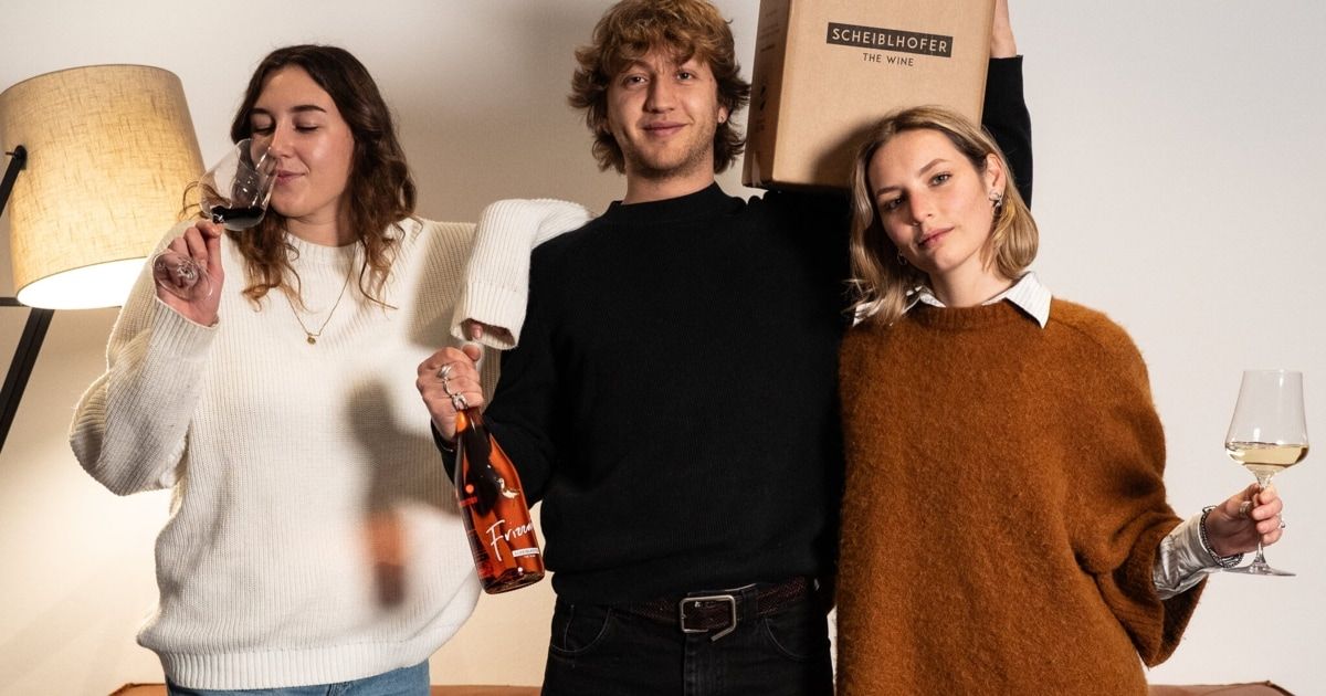 Drei Menschen stehen in einem Raum zusammen. Ein Mann in der Mitte hält eine Flasche Wein, während eine Frau links aus einem Glas trinkt. Eine Frau rechts lächelt. Sie stehen vor einer weißen Wand.