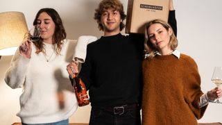 Drei Menschen stehen in einem Raum zusammen. Ein Mann in der Mitte hält eine Flasche Wein, während eine Frau links aus einem Glas trinkt. Eine Frau rechts lächelt. Sie stehen vor einer weißen Wand.