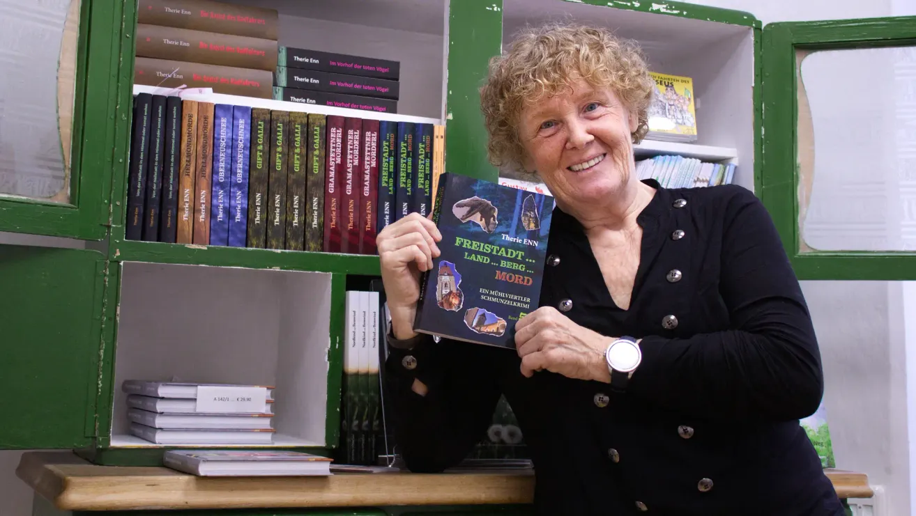 Eine ältere Frau lächelt und hält ein Buch mit dem Titel Freistadt Land...Berg...Mord vor einem mit Büchern gefüllten Bücherregal.