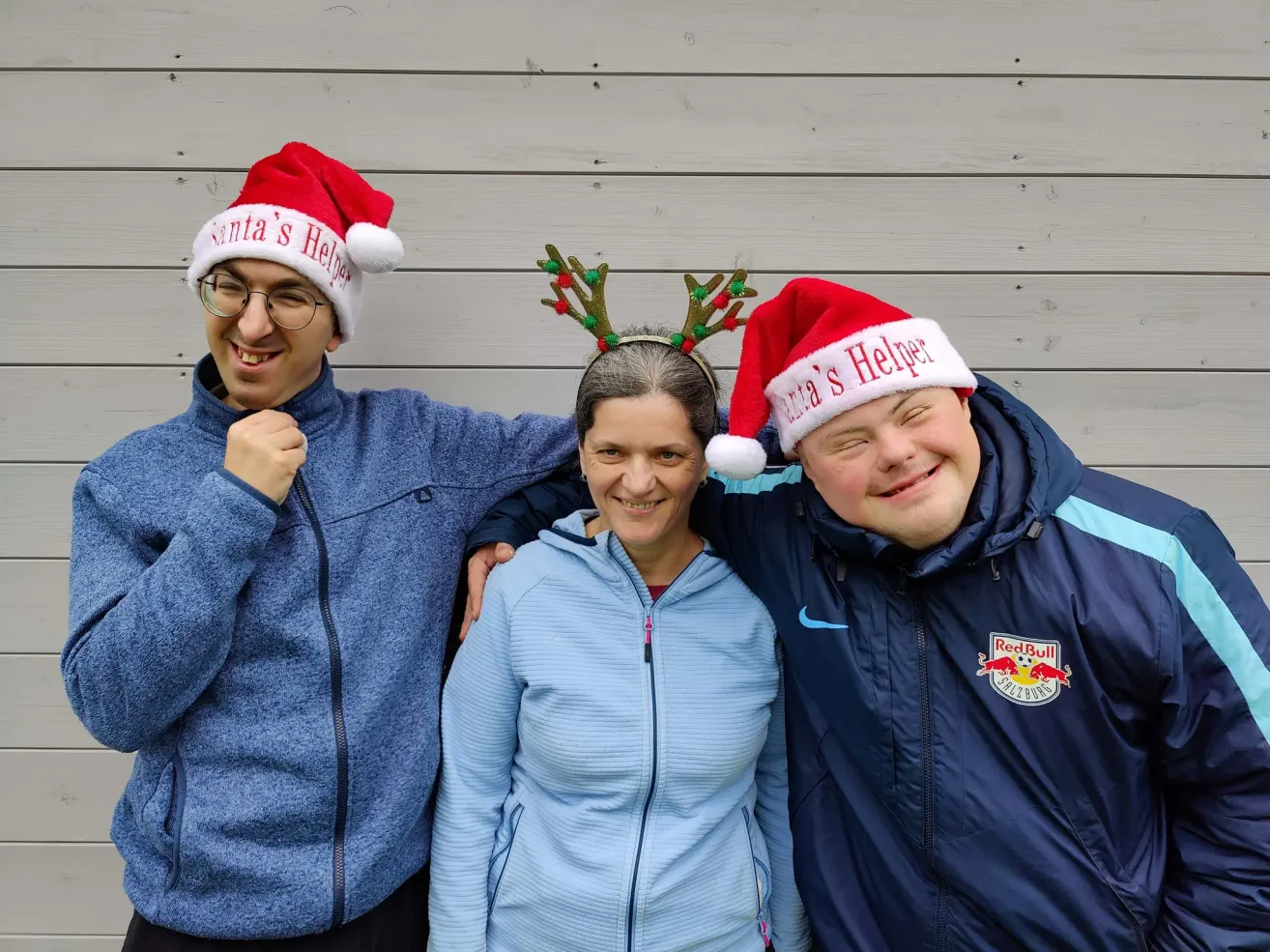 Drei Personen in Winterkleidung und Weihnachtsmützen, zwei mit Rentiergeweihen, lächeln für ein Foto. Der Hintergrund ist eine weiße Holzwand.