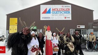 Eine Gruppe von Menschen in Kostümen posiert vor einem Gebäude, darunter zwei Mädchen in weißen Kostümen mit Körben, ein Mann in einem Weihnachtsmann-Kostüm und mehrere Personen in Teufelskostümen mit Hörnern.