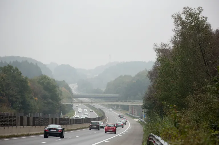 Eine neblige Autobahn mit mehreren fahrenden Autos, einer Brücke in der Ferne und Grünanlagen entlang der Seiten.