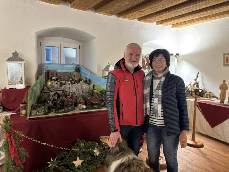 Zwei Personen stehen vor einer Weihnachtsdekoration. Ein Mann in einem roten Mantel hält einen Hund. Eine Frau trägt eine Brille und einen Schal.