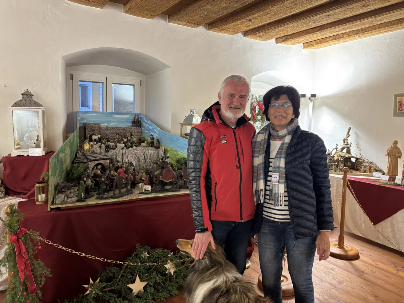 Zwei Personen stehen vor einer Weihnachtsdekoration. Ein Mann in einem roten Mantel hält einen Hund. Eine Frau trägt eine Brille und einen Schal.