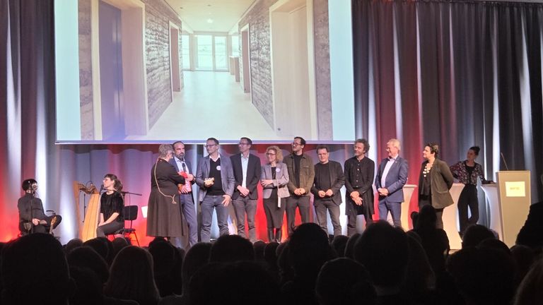 Eine Gruppe von Menschen steht auf einer Bühne mit einem Projektor dahinter. Sie scheinen eine Präsentation zu halten. Der Raum hat eine rot-weiße Wand mit Vorhängen an der Seite.