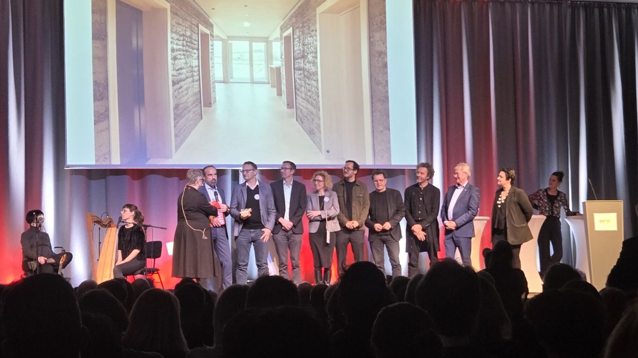 Eine Gruppe von Menschen steht auf einer Bühne mit einem Projektor dahinter. Sie scheinen eine Präsentation zu halten. Der Raum hat eine rot-weiße Wand mit Vorhängen an der Seite.