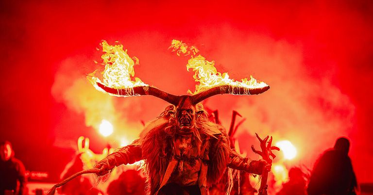 Eine gehörnte Figur steht in einer Menschenmenge und hält brennende Fackeln an ihren Hörnern, vor einem Hintergrund aus roten Lichtern und Feuer.