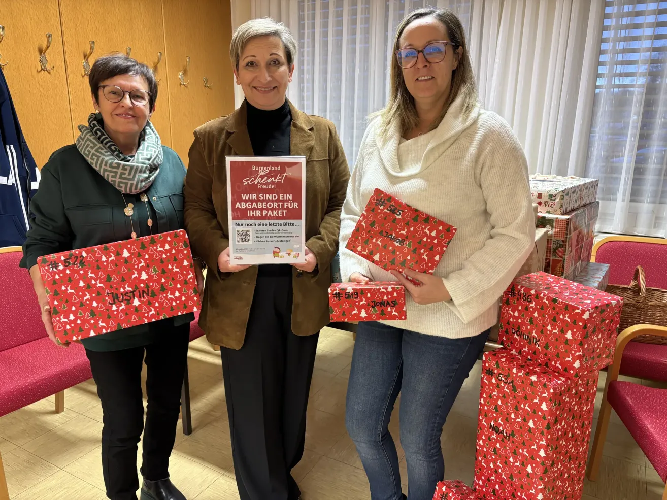 Drei Frauen stehen in einem Raum und halten Geschenkboxen. Eine Frau in der Mitte hält ein Schild mit der Aufschrift 'Wir sind ein Geschenkpaket für Ihr Paket'. Die Frau rechts trägt eine Brille und hält eine Geschenkbox. Hinter ihnen stehen weitere Geschenkboxen.