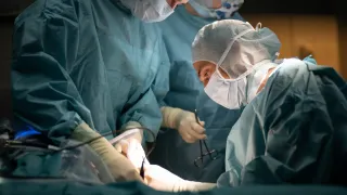 Surgeons in blue scrubs and masks work in an operating room, one holding a scalpel. The surgeon in front is closely examining a patient.