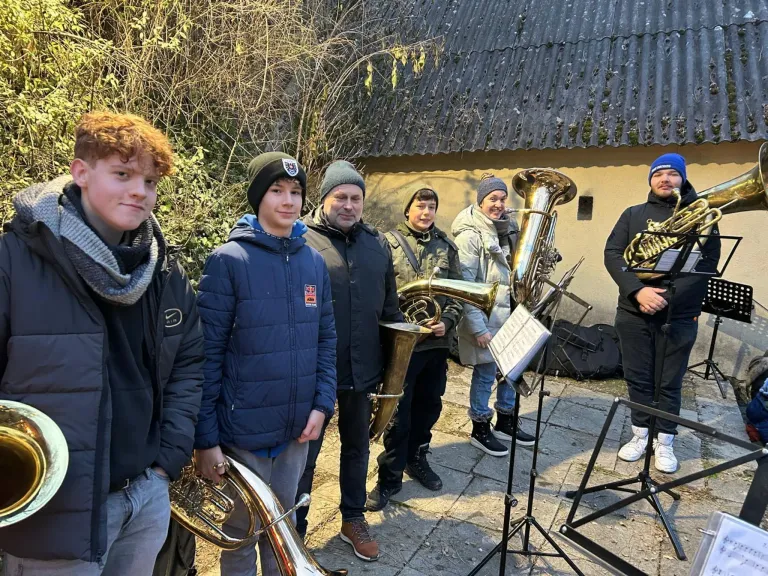 Eine Gruppe von Menschen in Winterkleidung steht vor einem Gebäude und hält Blechblasinstrumente. Sie scheinen eine Band zu sein, die sich möglicherweise auf eine Aufführung vorbereitet.
