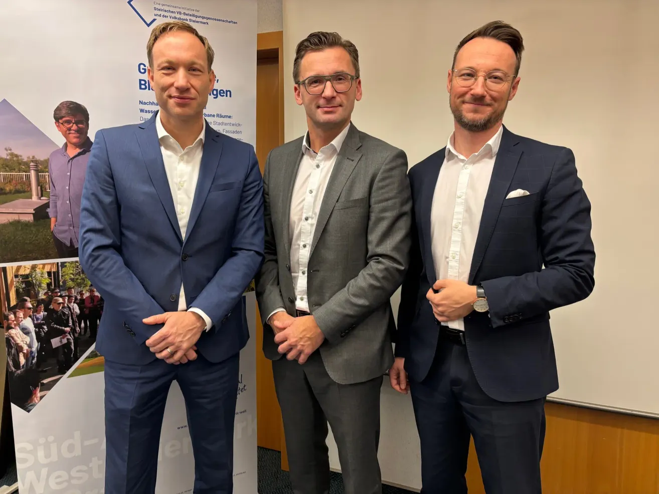 Drei Männer in Anzügen und mit Brillen stehen zusammen, wahrscheinlich in einer Besprechung oder einem professionellen Umfeld. Der Mann auf der linken Seite trägt einen blauen Anzug, der Mann in der Mitte einen grauen Anzug und der Mann auf der rechten Seite einen marineblauen Anzug. Sie scheinen zu lächeln und posieren für ein Foto.