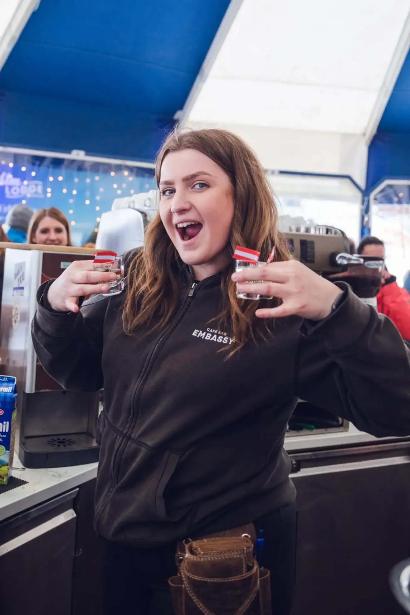 Eine Frau in einer schwarzen Jacke hält zwei Getränke in einem Café. Im Hintergrund ist eine Person in einer roten Jacke zu sehen.