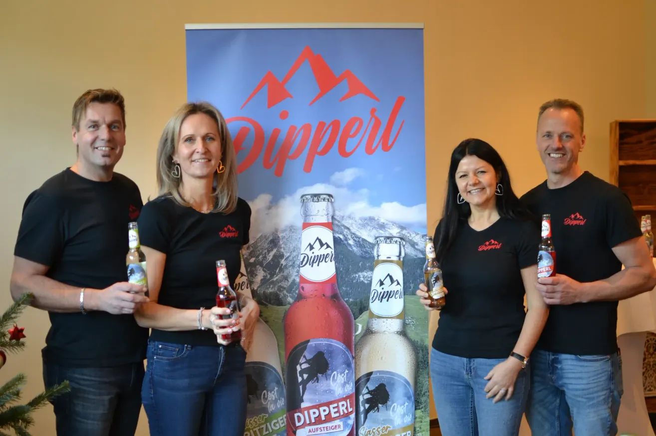 Vier Personen stehen neben einem Banner mit Dipperl-Flaschen in den Händen. Das Banner zeigt eine Bergkette und das Wort Dipperl. Die Leute lächeln.