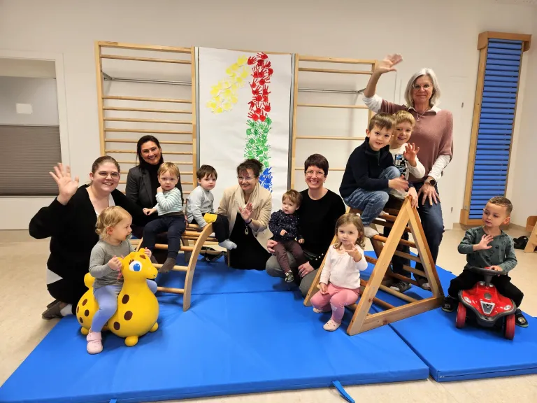 Eine Gruppe von Erwachsenen und Kindern posiert für ein Foto in einem Spielzimmer mit bunten Strukturen und einem Regenbogen-Hintergrund. Einige Kinder sitzen auf Holzleitern, während andere auf dem Boden sitzen.