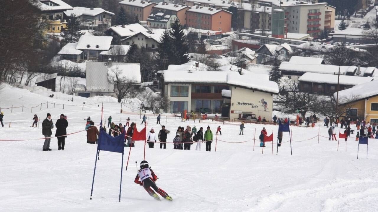 Ein Skirennen findet statt, ein Skifahrer navigiert durch verschneite Tore. Zuschauer stehen hinter Seilen und beobachten das Ereignis. Die Gebäude im Hintergrund sind mit Schnee bedeckt.
