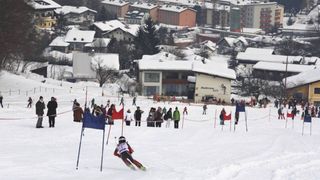 A ski race is in progress with a skier navigating through snowy gates. Spectators stand behind ropes, watching the event. Buildings in the background are blanketed with snow.
