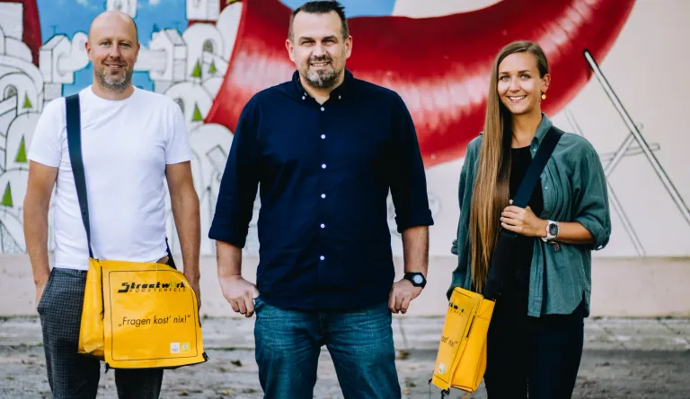 Drei Personen stehen vor einer Graffiti-Wand. Ein Mann in einem dunklen Hemd, eine Frau mit einer gelben Tasche und eine weitere Person in einem weißen T-Shirt. Sie alle lächeln.