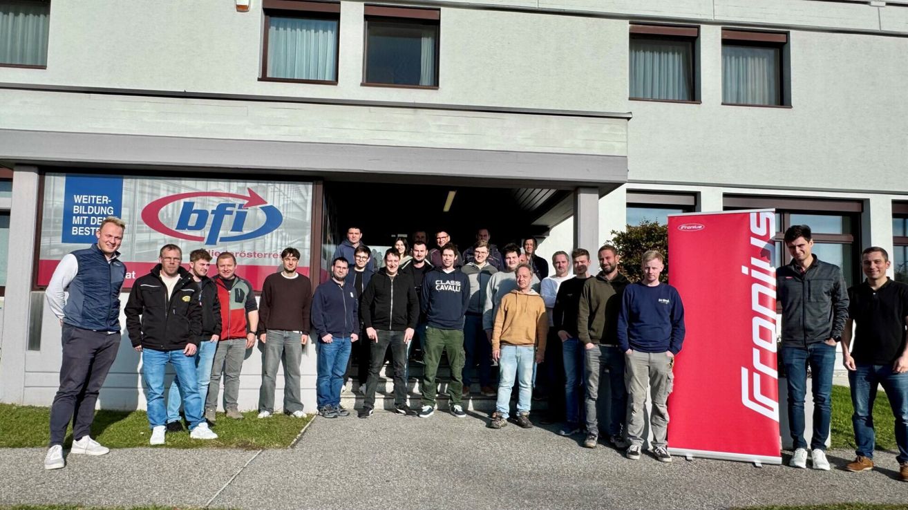 Eine Gruppe von Menschen in Jacken und Turnschuhen steht vor einem Gebäude. Einige tragen Brillen. Ein Banner ist an der Wand hinter ihnen angebracht.