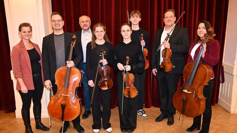 Sechs Musiker posieren mit ihren Instrumenten für ein Foto. Zwei Männer halten Cellos, zwei Frauen halten Violinen und zwei Männer halten Violinen. Sie stehen vor einem roten Vorhang.