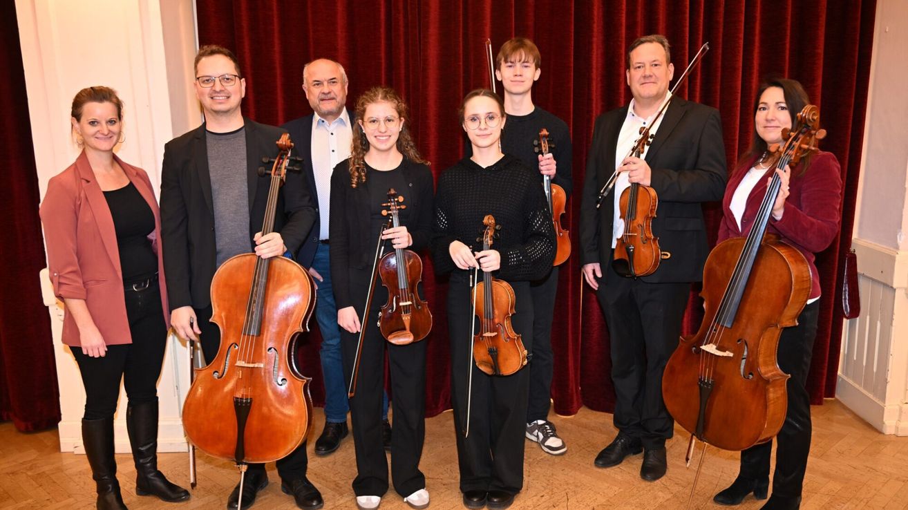 Sechs Musiker posieren mit ihren Instrumenten für ein Foto. Zwei Männer halten Cellos, zwei Frauen halten Violinen und zwei Männer halten Violinen. Sie stehen vor einem roten Vorhang.