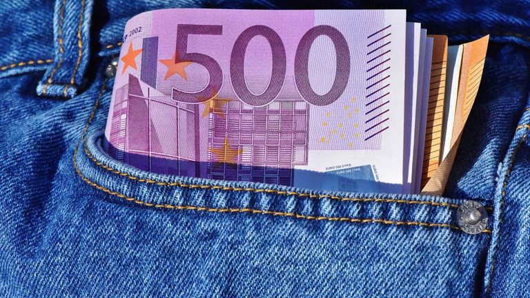 Eine Nahaufnahme einer gefalteten 500-Euro-Banknote, sichtbar von hinten, in einer blauen Jeanstasche verstaut. Die Banknote hat ein lila und weißes Design mit Sternen und geometrischen Mustern.