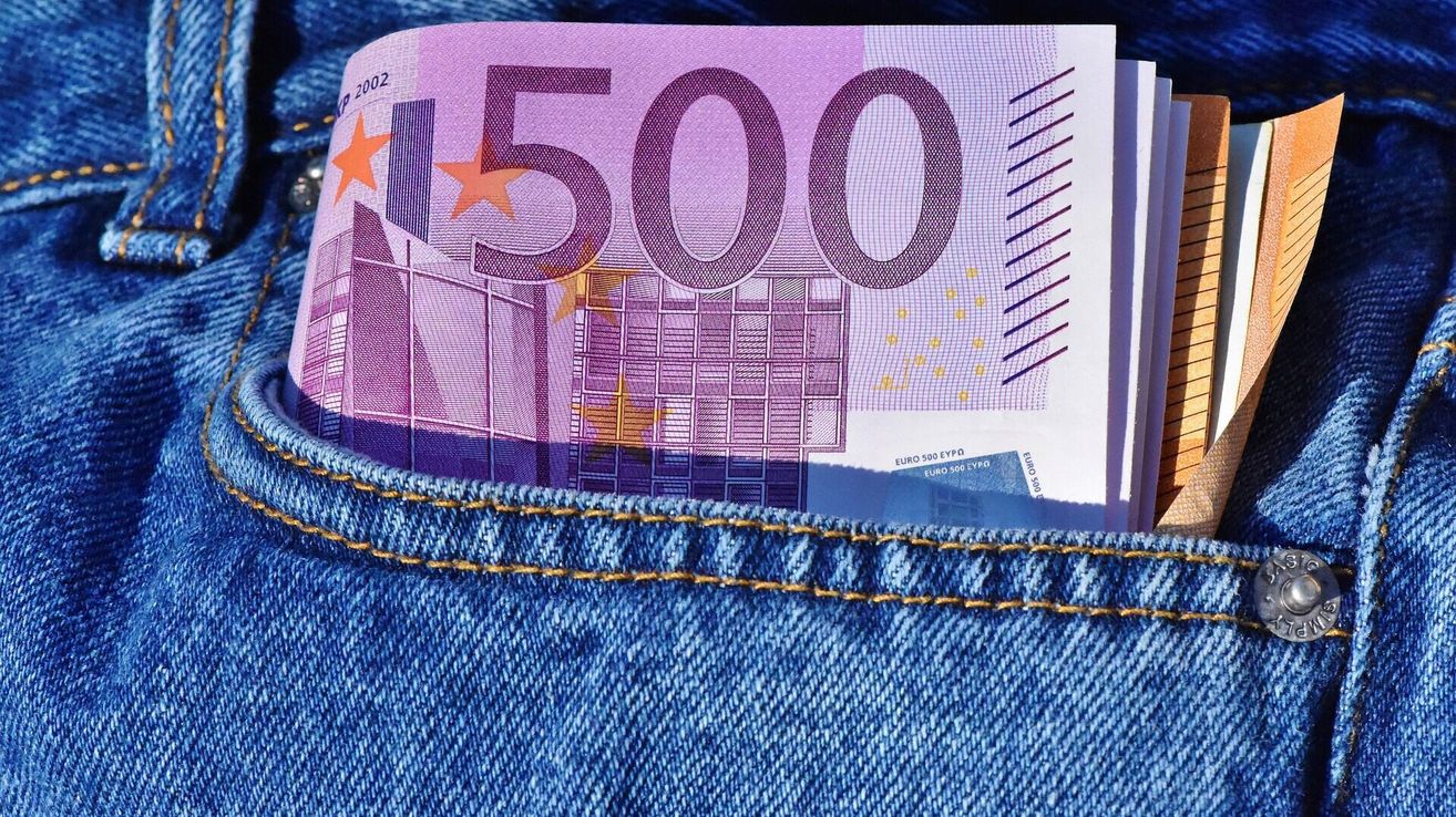 Eine Nahaufnahme einer gefalteten 500-Euro-Banknote, sichtbar von hinten, in einer blauen Jeanstasche verstaut. Die Banknote hat ein lila und weißes Design mit Sternen und geometrischen Mustern.