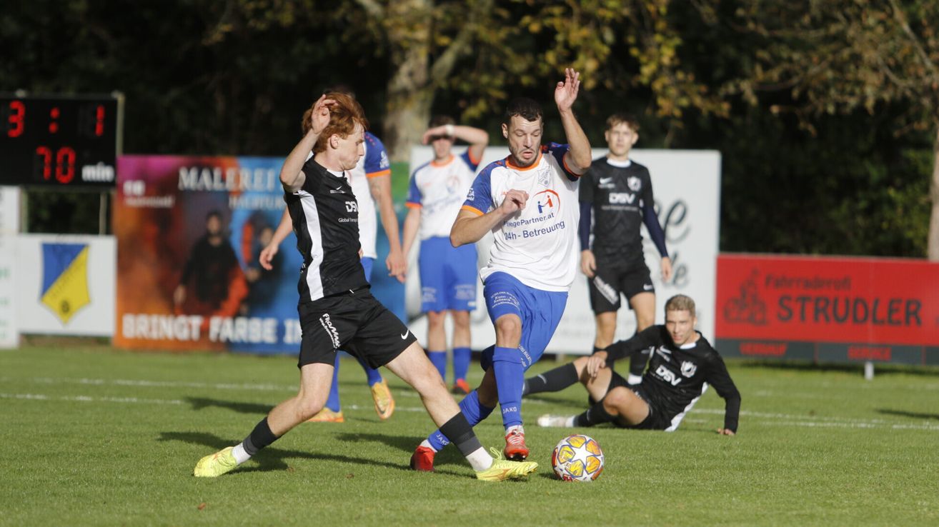 Ein Fußballspiel mit Spielern in verschiedenen Trikots. Ein Spieler hebt die Hand, ein anderer liegt auf dem Boden. Ein Spieler in Blau und Weiß verfolgt den Ball.