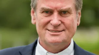 Ein Mann in einem dunkelblauen Anzug mit einem weißen Hemd mit Kragen, lächelt für ein Foto, mit einem unscharfen grünen Hintergrund.