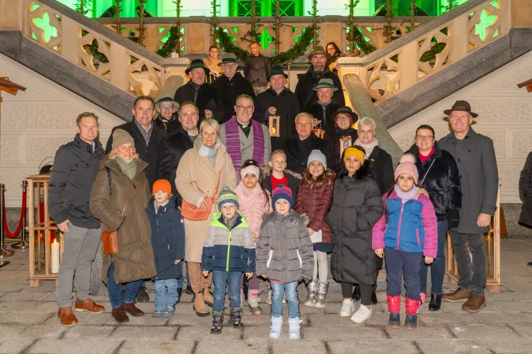 Eine Gruppe von Menschen, darunter Kinder, steht auf einer Treppe mit verzierten Geländern. Sie tragen alle Winterkleidung. Einige halten Laternen. Dahinter stehen weitere Menschen in der Nähe der Geländer.