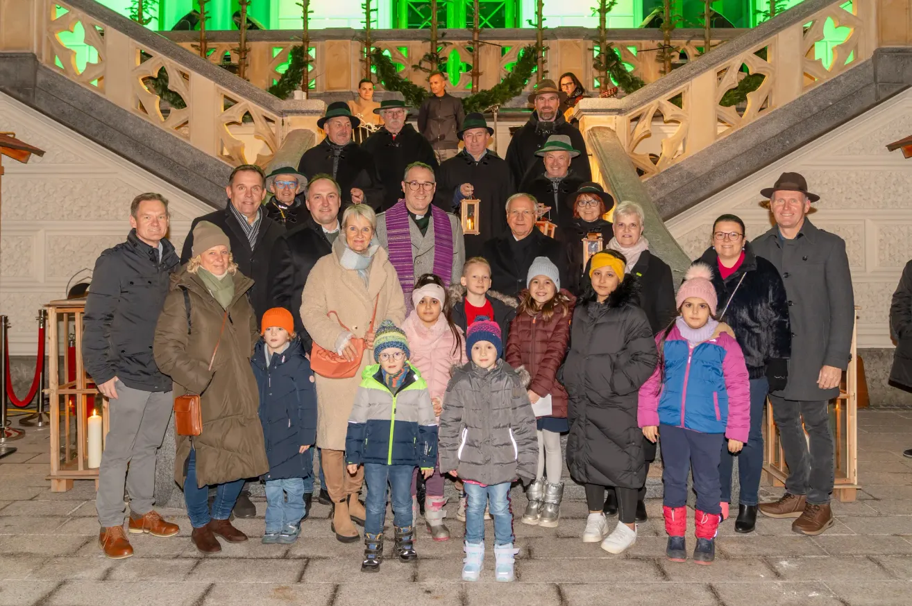 Eine Gruppe von Menschen, darunter Kinder, steht auf einer Treppe mit verzierten Geländern. Sie tragen alle Winterkleidung. Einige halten Laternen. Dahinter stehen weitere Menschen in der Nähe der Geländer.
