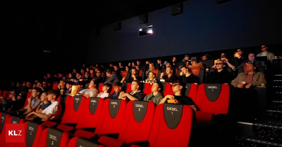 Publikum sitzt auf roten Stühlen in einem Kinosaal. Die Stühle sind mit 'Diesel 1' bis 'Diesel 5' beschriftet. Die Leute halten Snacks in den Händen und tragen Brillen. Einige haben die Augen geschlossen, wahrscheinlich beim Anschauen des Films. Das Kino ist dunkel beleuchtet.